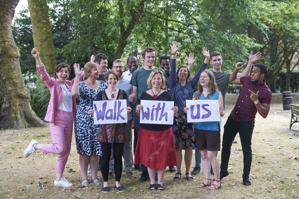 A group of people in a park, smiling and holding their hands in the air. The three women at the front a each holding a piece of paper with a word, spelling out 'Walk with us'.