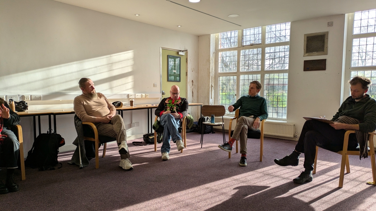 A group of five people sit in a semi-circle in a sunny room with tall windows. One man, the group leader, is talking, while the others look like they are listening or taking notes.