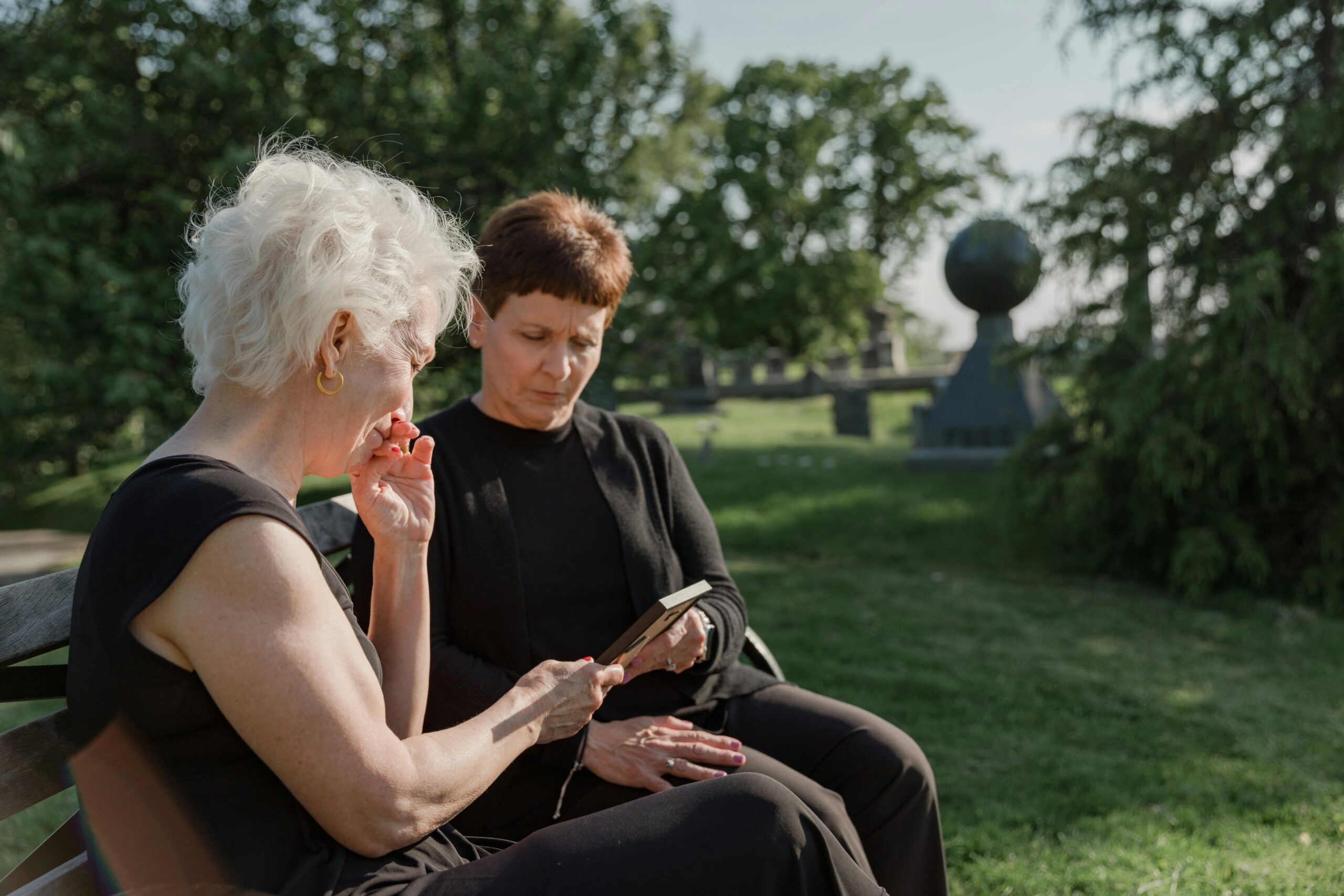 Two white women are sat on a bench in a park. They are looking at a photograph in a frame that the older woman is holding.