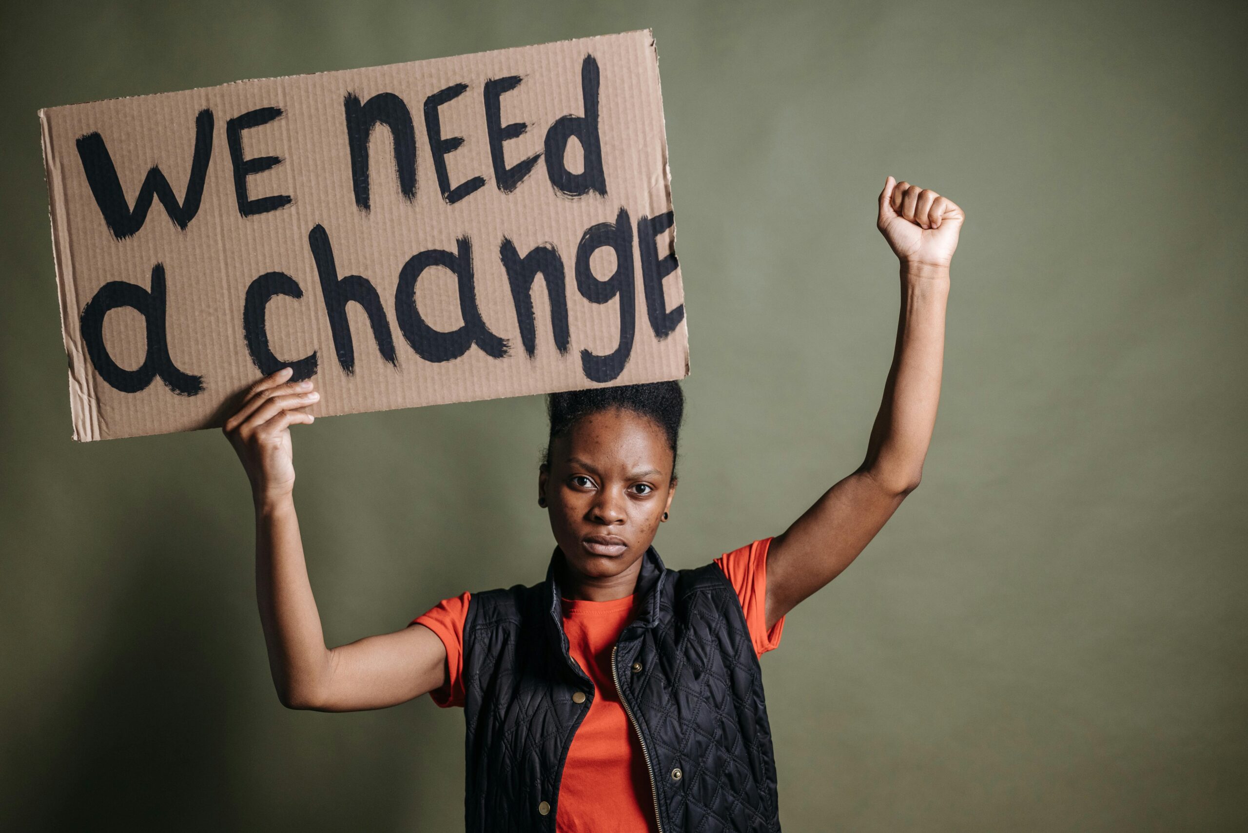 A black woman is standing holding a placard which reads "We need a change". She punches the air with her other hand.
