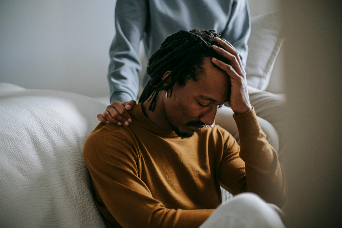 A man sits in front of a bed with his head in his hands and his eyes closed. Another person - whose face we cannot see - is sitting on the bed and has their hand on his shoulder.