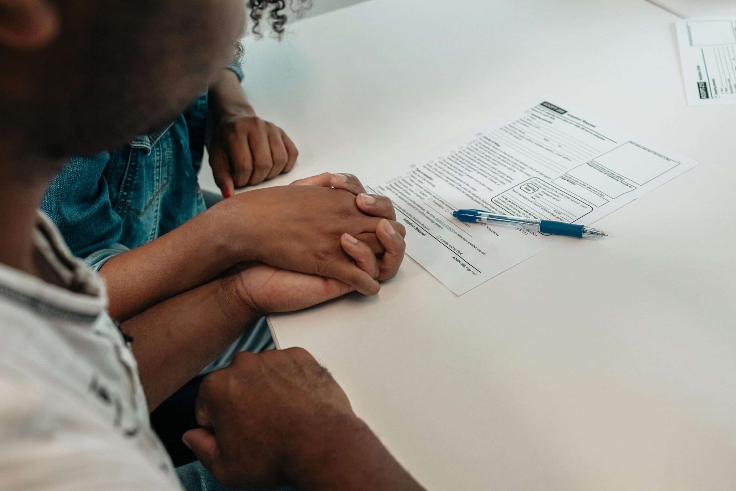 A black couple are sat at a table holding hands. On the table is a paper form and a pen.