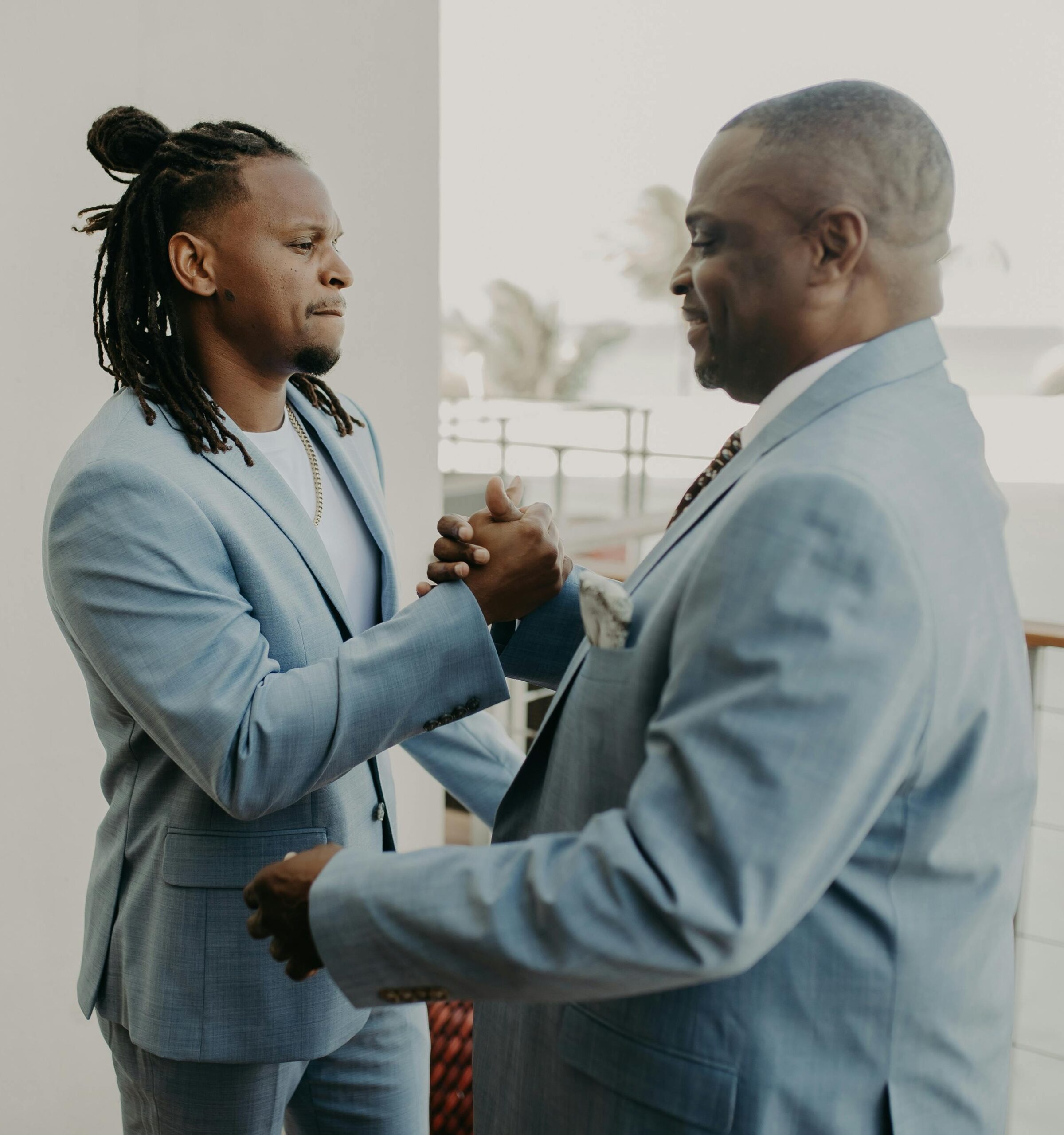 An older bald black man and a younger black man with dreadlocks are wearing light blue suits and engaging in a bro handshake.