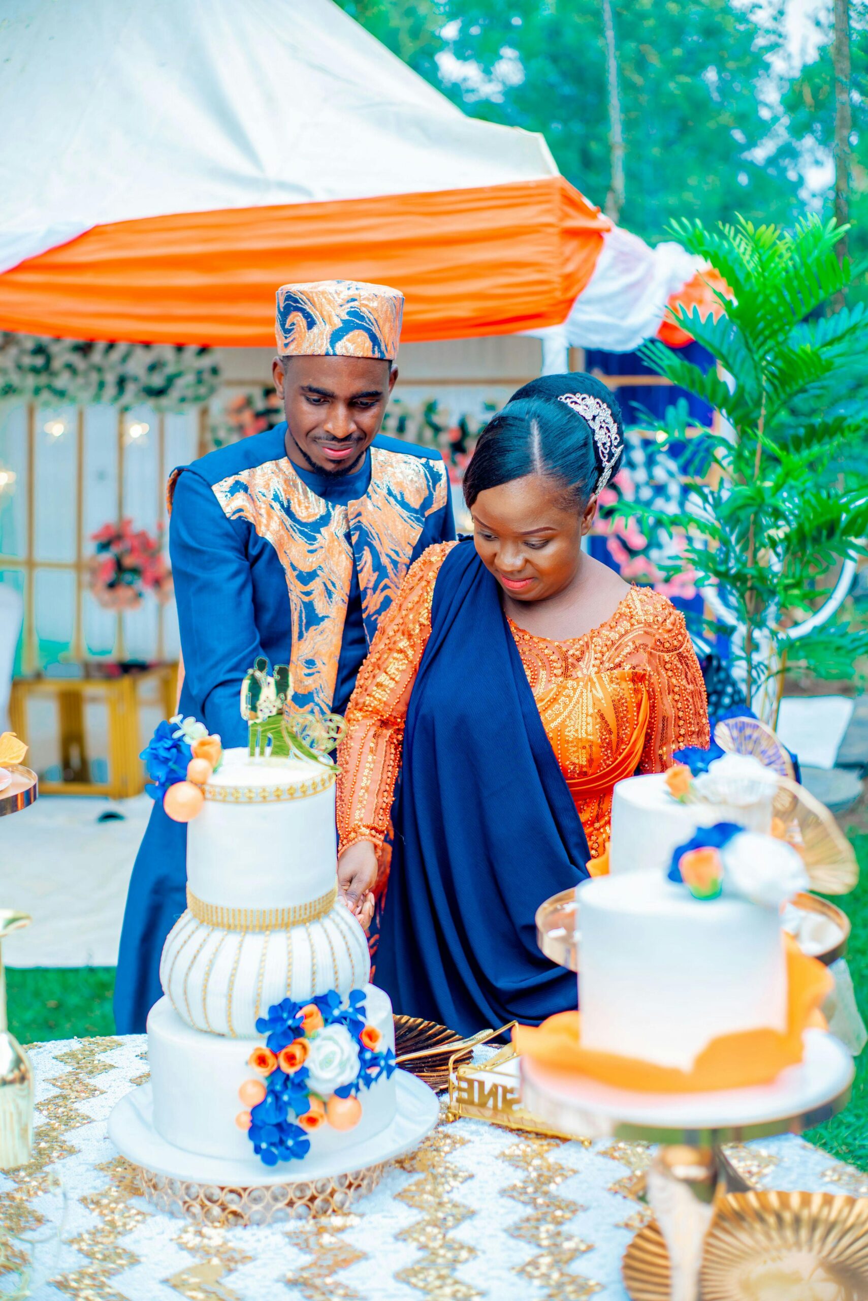 A black man and woman are stood outside wearing vibrant blue and orange traditional African clothing. They are cutting a wedding cake together.