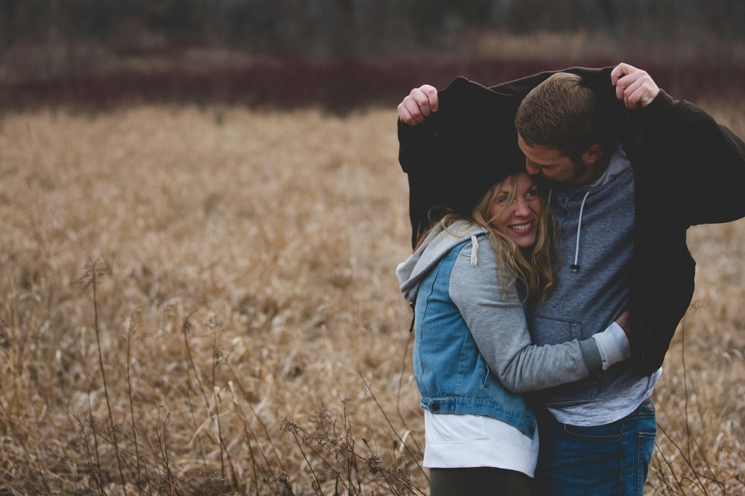 A young, white man and woman are standing in a brown field. The man is holding his jacket over their heads as if to protect from rain. The woman is smiling.