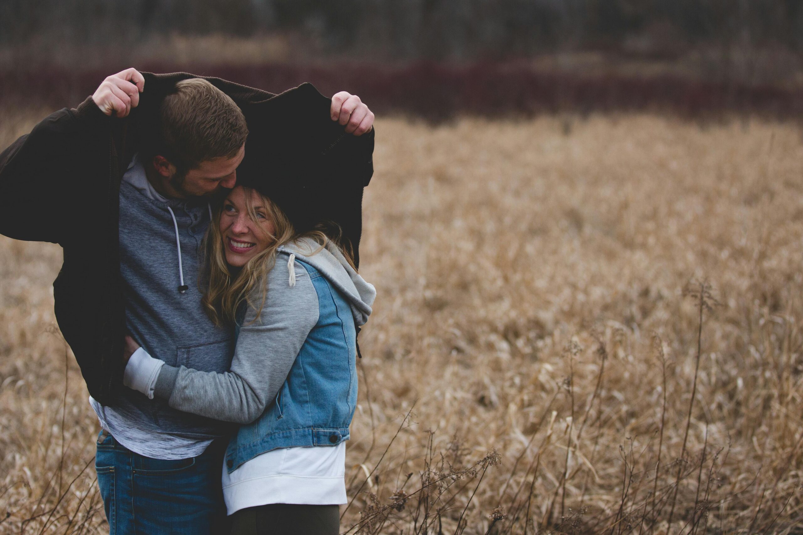 A young, white man and woman are standing in a brown field. The man is holding his jacket over their heads as if to protect from rain. The woman is smiling.