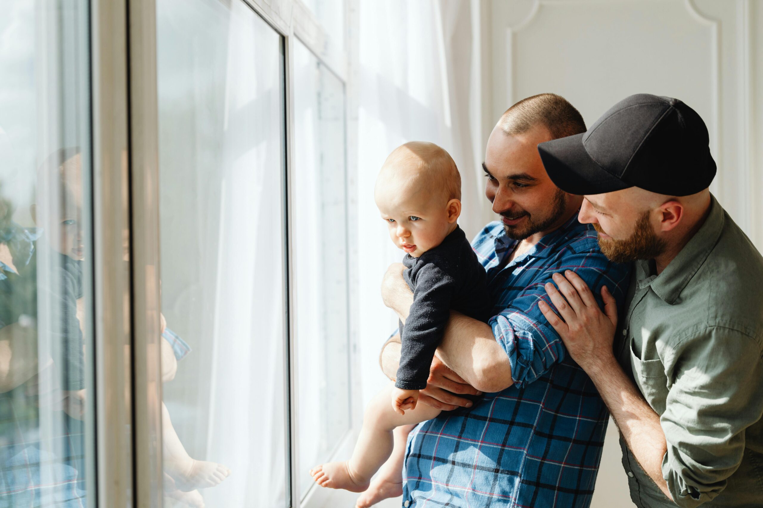 A white male couple are stood by a window. One is holding a baby. The other is stood slightly behind with his hand affectionately on his partner's arm.
